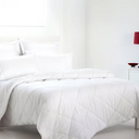 A white quilted comforter with matching pillows and a folded back white duvet on a bed in a bright, minimalist bedroom.